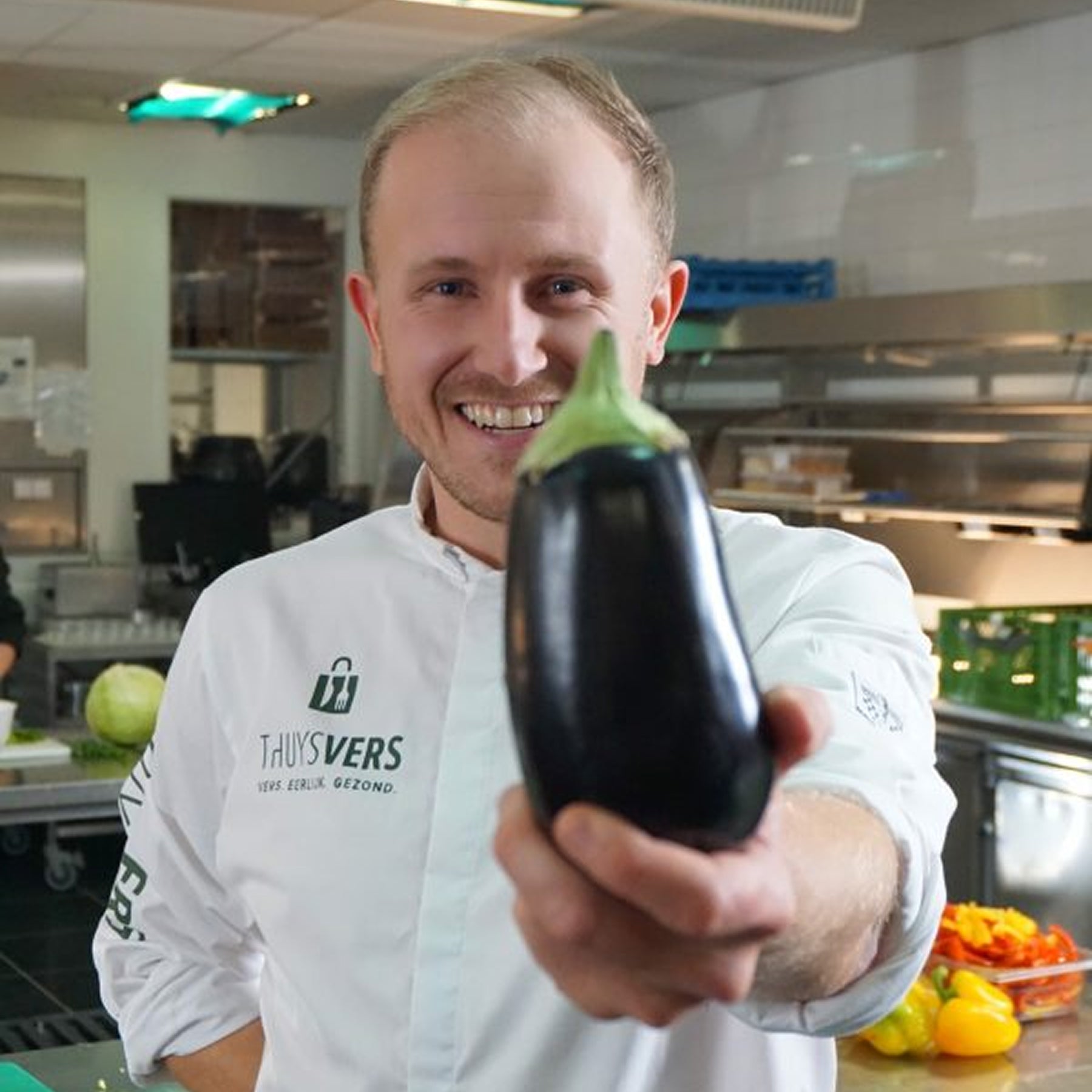 Kok in professionele keuken houdt een glanzende aubergine vast met een brede glimlach, omringd door verse groenten en apparatuur.